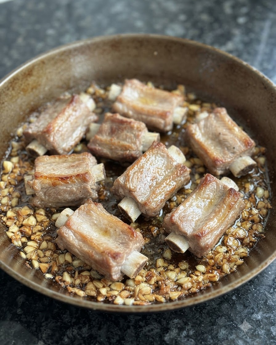 Costillas de cerdo escaldadas dorándose en una sartén rodeadas de ajo frito y aceite.