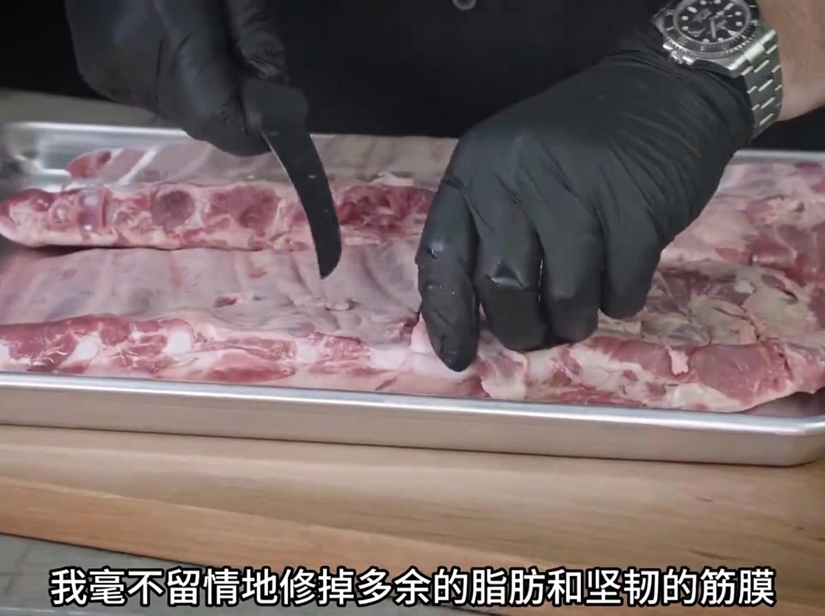 Un par de manos con guantes usando un cuchillo para recortar la grasa de las costillas de cerdo crudas en una bandeja de metal.