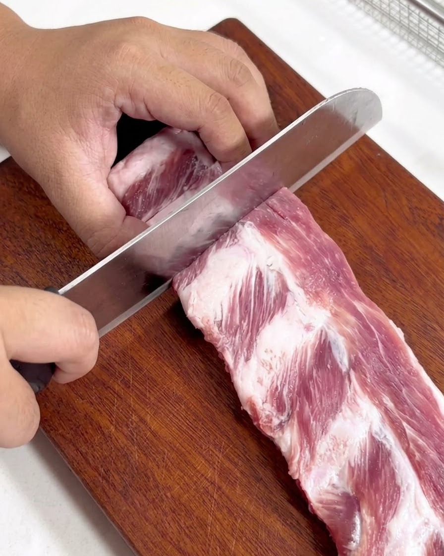 Un costillar de cerdo crudo cortándose en piezas individuales más pequeñas sobre una tabla de cortar de madera.