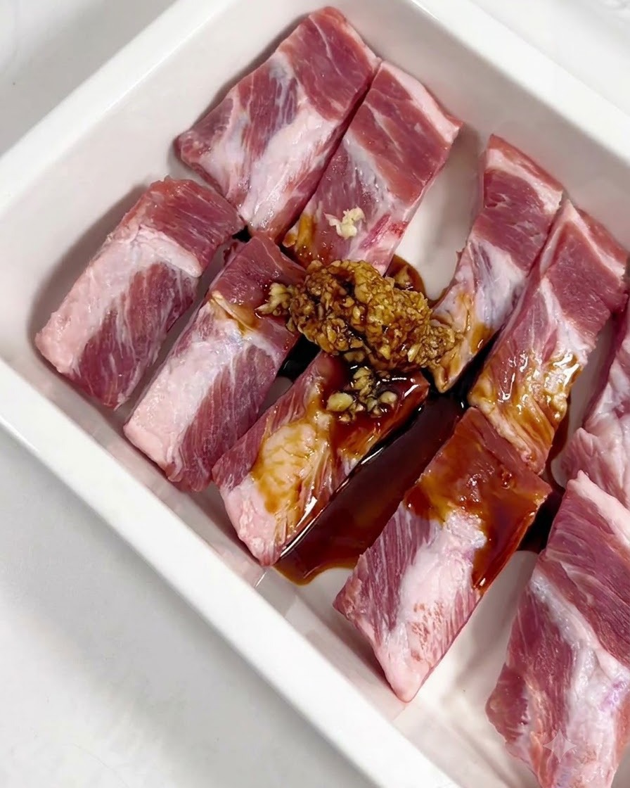 Ajo picado, salsa de soja, salsa de ostras, miel y condimentos en polvo vertidos sobre costillas de cerdo crudas en una bandeja blanca.