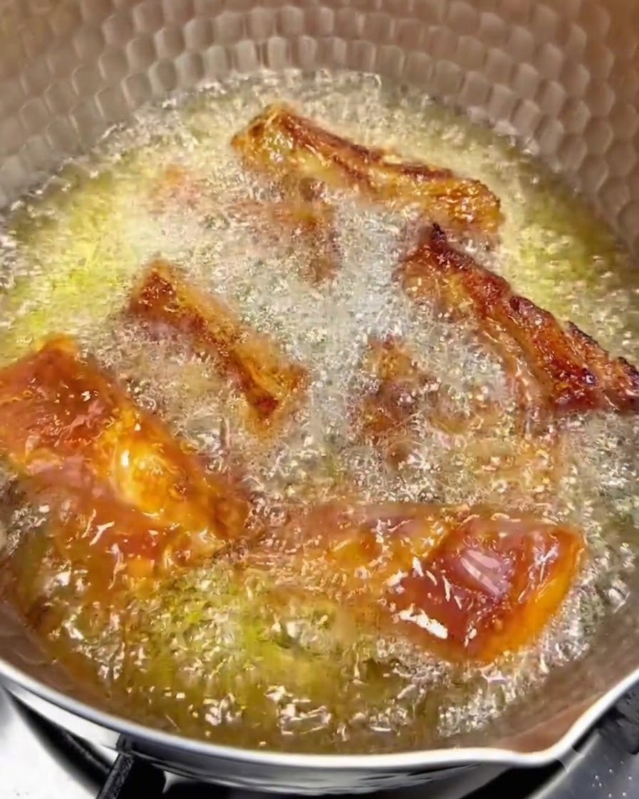 Costillas de cerdo friéndose en aceite caliente a fuego bajo con superficies doradas visibles.