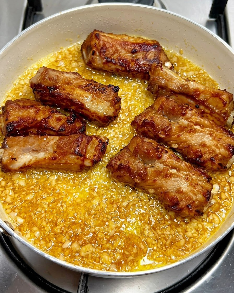 Costillas de cerdo fritas doradas dispuestas ordenadamente dentro de una sartén llena de salsa de mantequilla con ajo burbujeante.