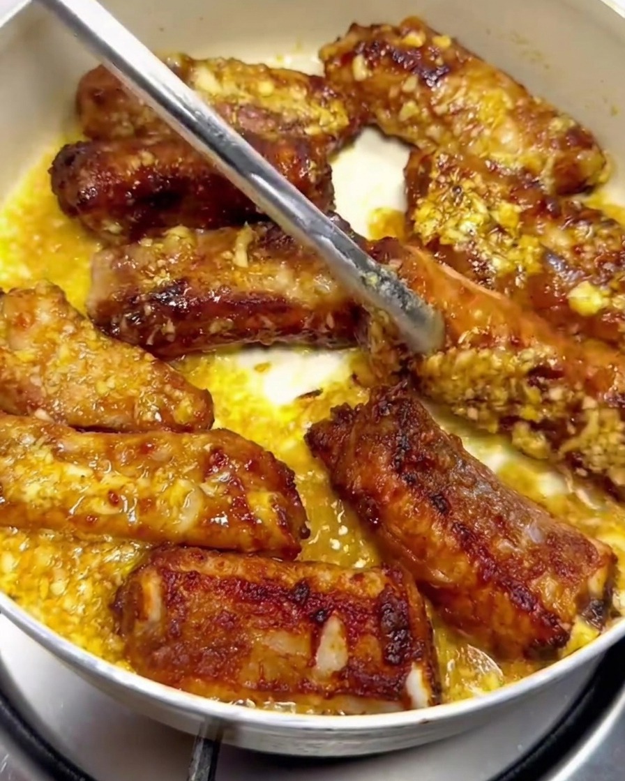 Costillas de cerdo girándose con pinzas de metal dentro de una sartén, cubiertas con salsa de mantequilla con ajo.