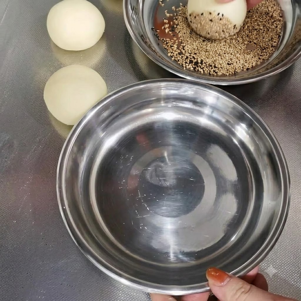 Una bola de masa siendo rebozada con semillas de sésamo en un bol metálico junto a otras bolas de masa preparadas.