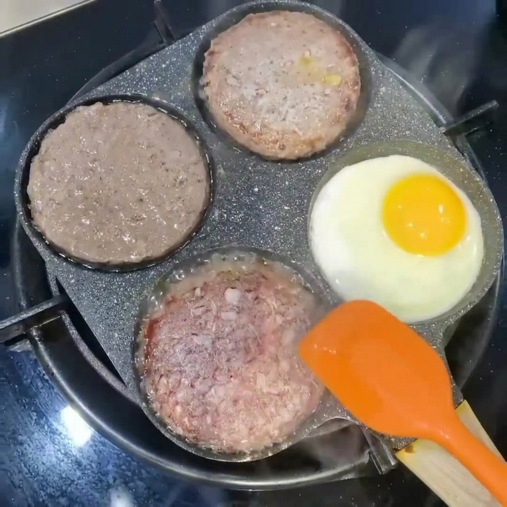 Hamburguesas siendo volteadas con una espátula naranja junto a un huevo friéndose en una sección de la sartén.