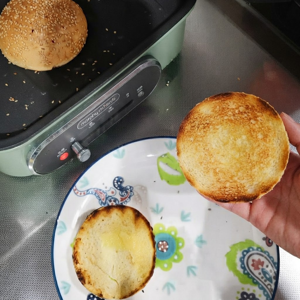 Mitades de pan de hamburguesa tostadas en un plato junto a una sartén con un panecillo entero tostándose.
