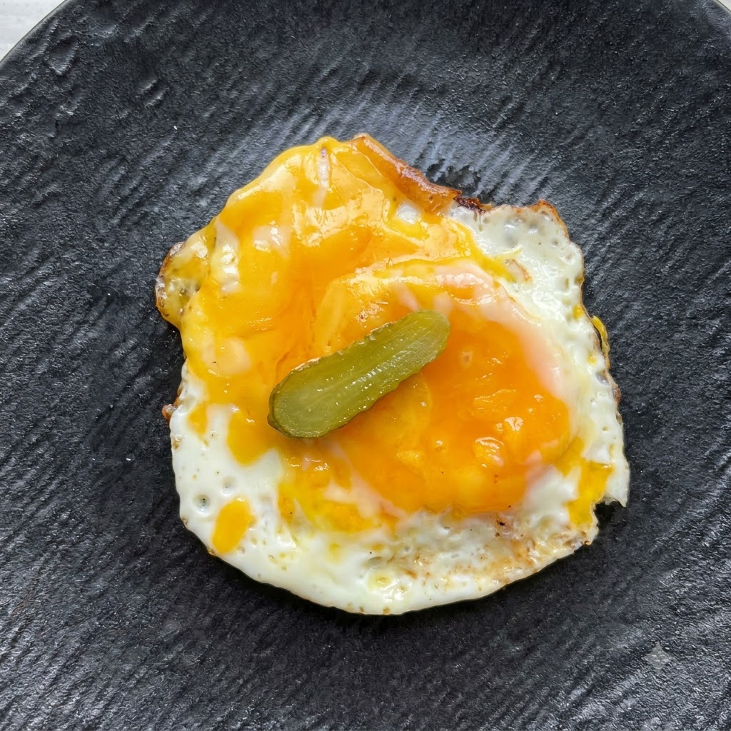 Una sola rodaja de pepinillo colocada en el centro sobre el huevo frito y el queso.