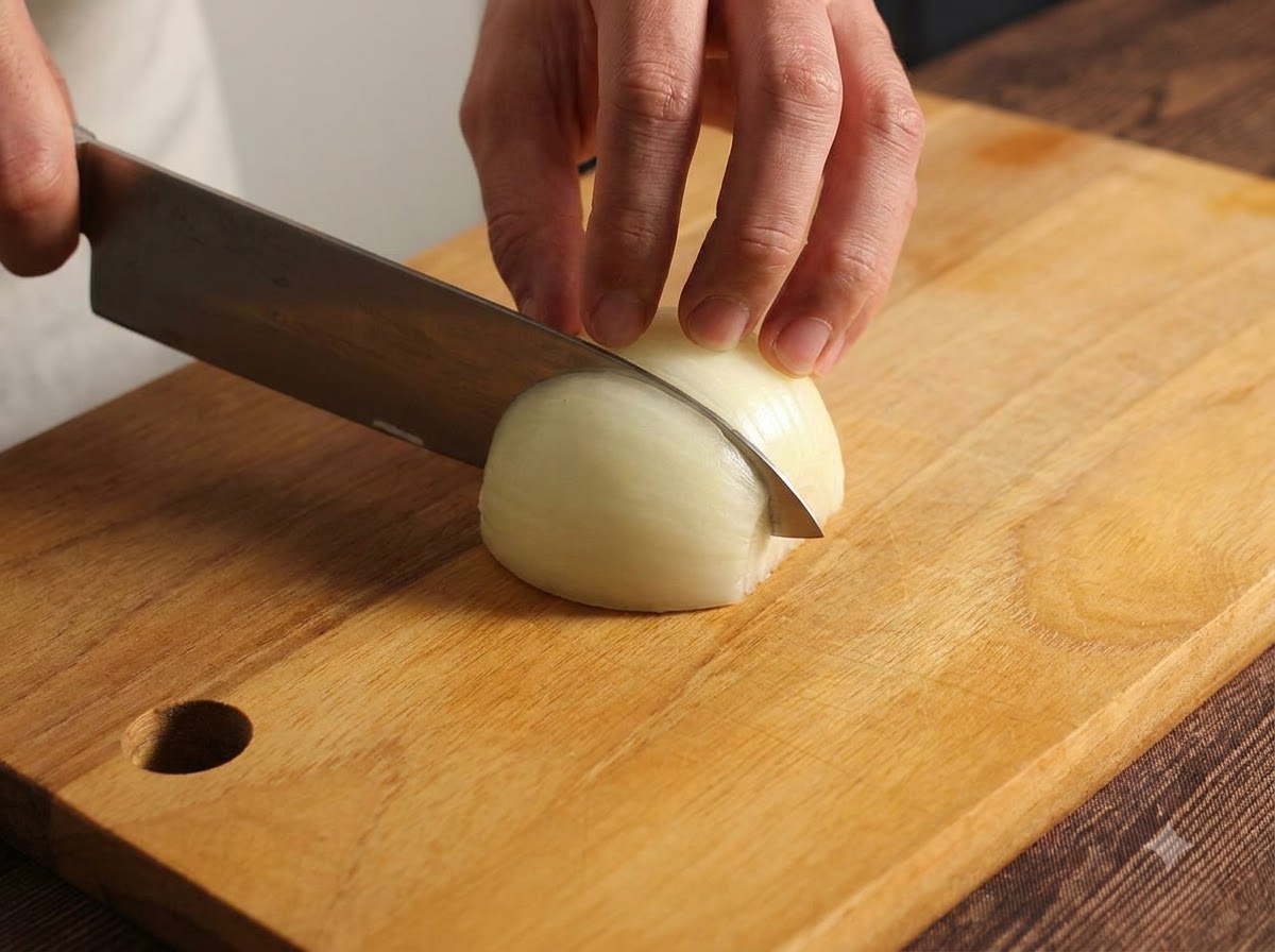 Una cebolla siendo cuidadosamente picada en una tabla de cortar de madera usando un cuchillo de cocina.