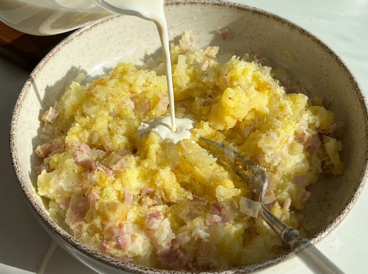 Crema de leche siendo vertida en un bol que contiene puré de papas y jamón picado.