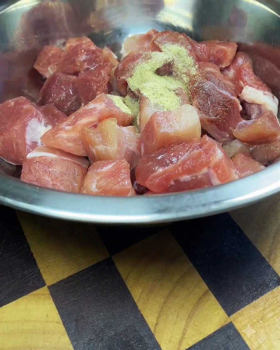 Cubos de cerdo crudos recién cortados dentro de un bol de mezcla de acero inoxidable espolvoreados con condimentos y fécula de maíz.