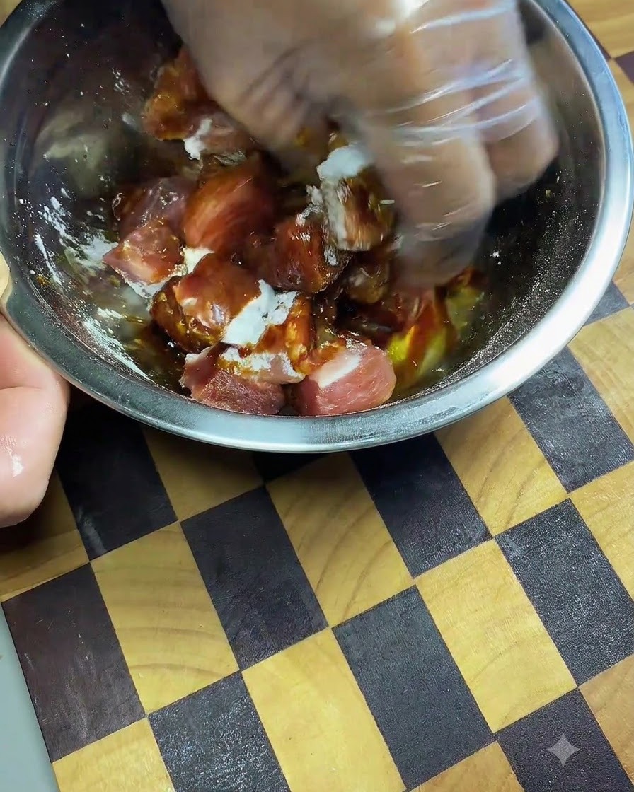 Una mano con guante amasando y masajeando salsa de soja oscura y especias en cubos de cerdo dentro de un bol de metal.