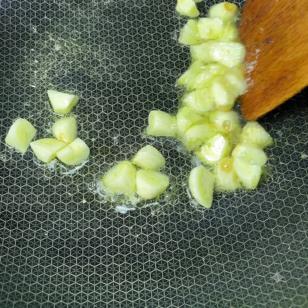 Dientes de ajo frescos picados chisporroteando en aceite caliente dentro de un wok siendo revueltos con una espátula de madera.