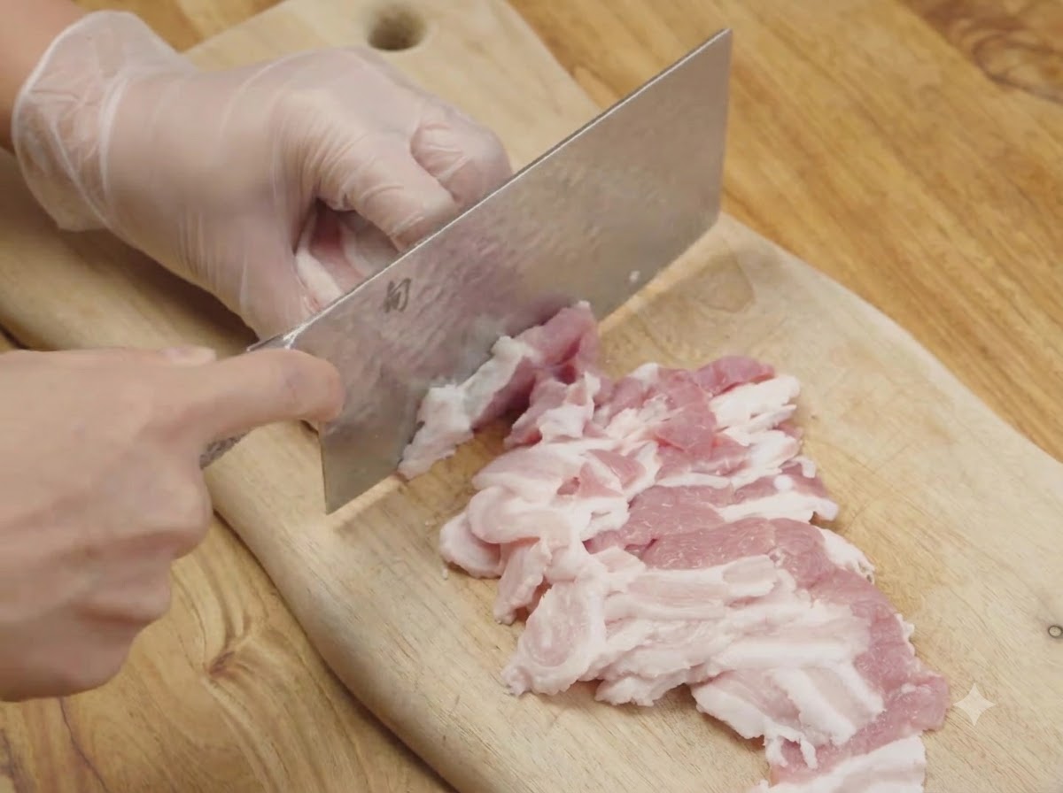 Panceta de cerdo cruda siendo cortada en tiras finas sobre una tabla de cortar de madera usando un cuchillo de cocina.