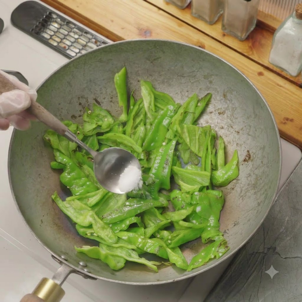 Una cucharada de sal siendo añadida a los pimientos verdes cortados que se tuestan en seco en un wok.