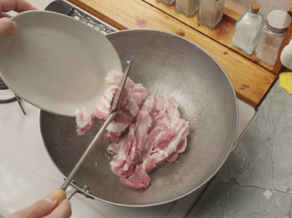 Panceta de cerdo cruda cortada siendo vertida desde un plato blanco pequeño a un wok caliente.