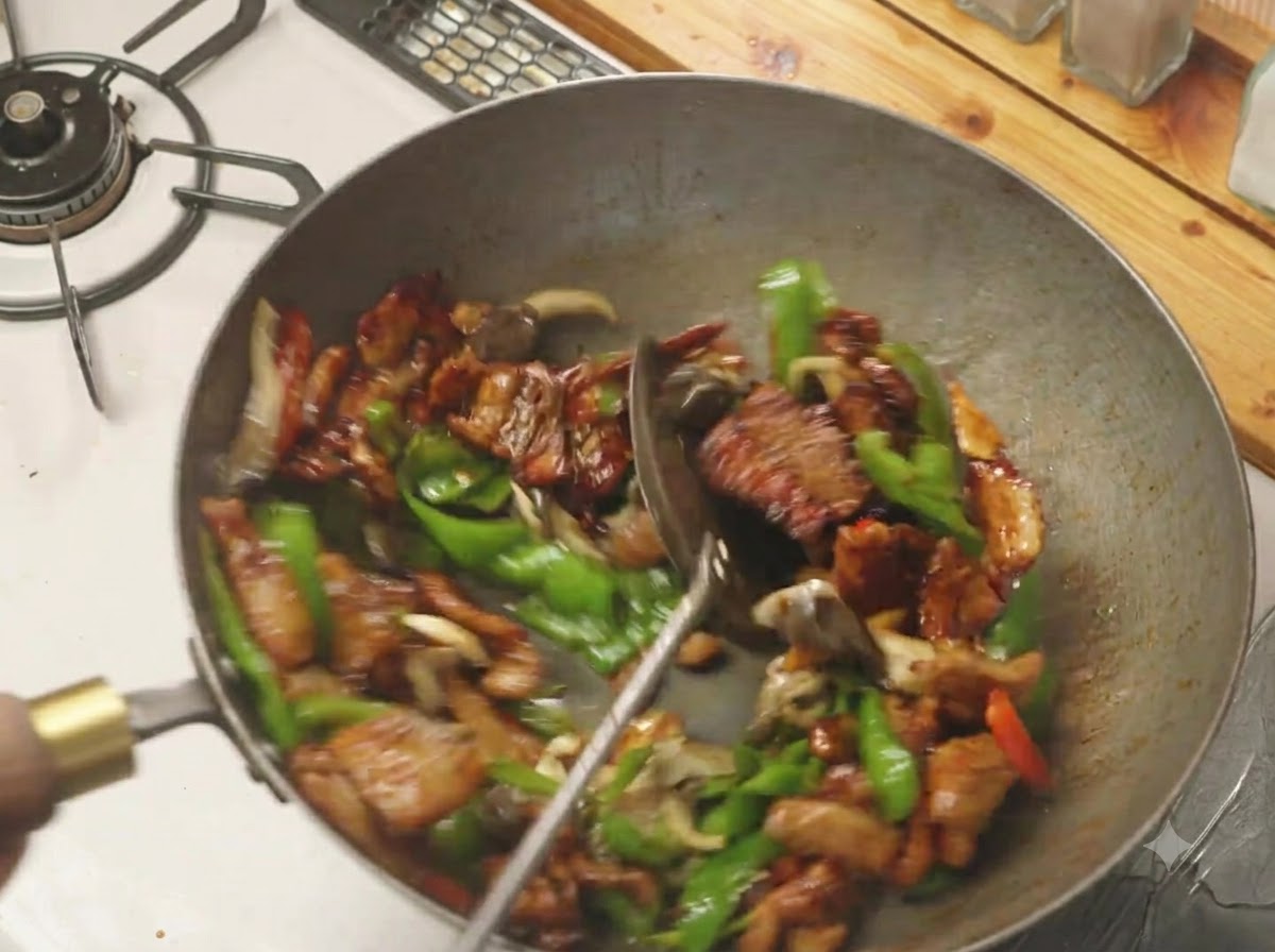 Una espátula mezclando pimientos verdes, setas y panceta de cerdo en un wok caliente.
