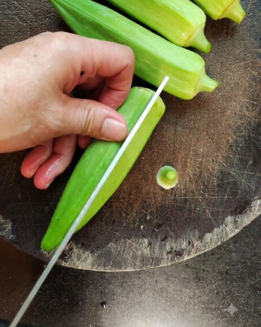 Un primer plano de un cuchillo dividiendo una okra verde fresca a lo largo en una tabla de cortar oscura.