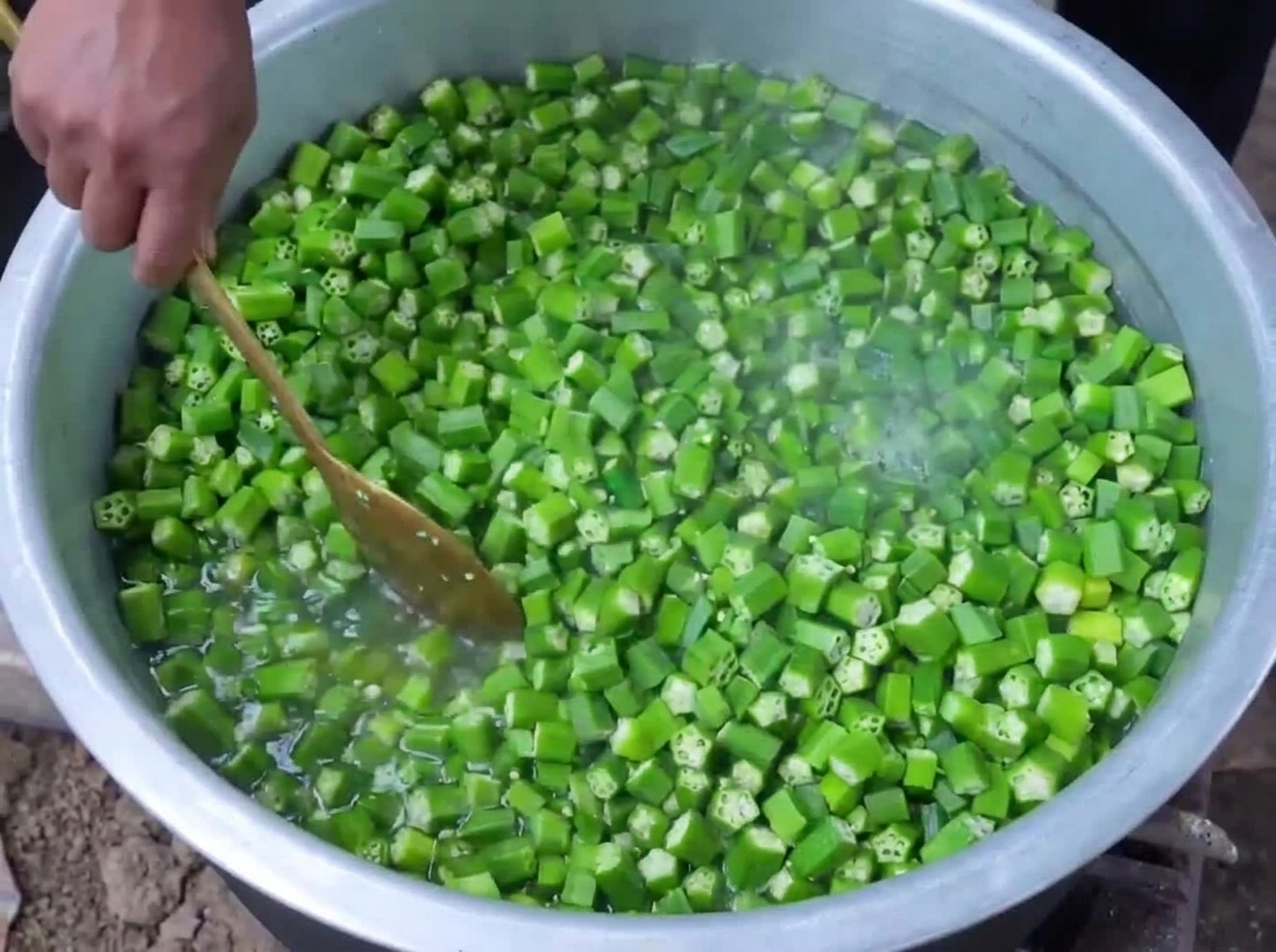 Okra verde picada siendo blanqueada en agua hirviendo dentro de una olla grande mientras se revuelve con una cuchara de madera.