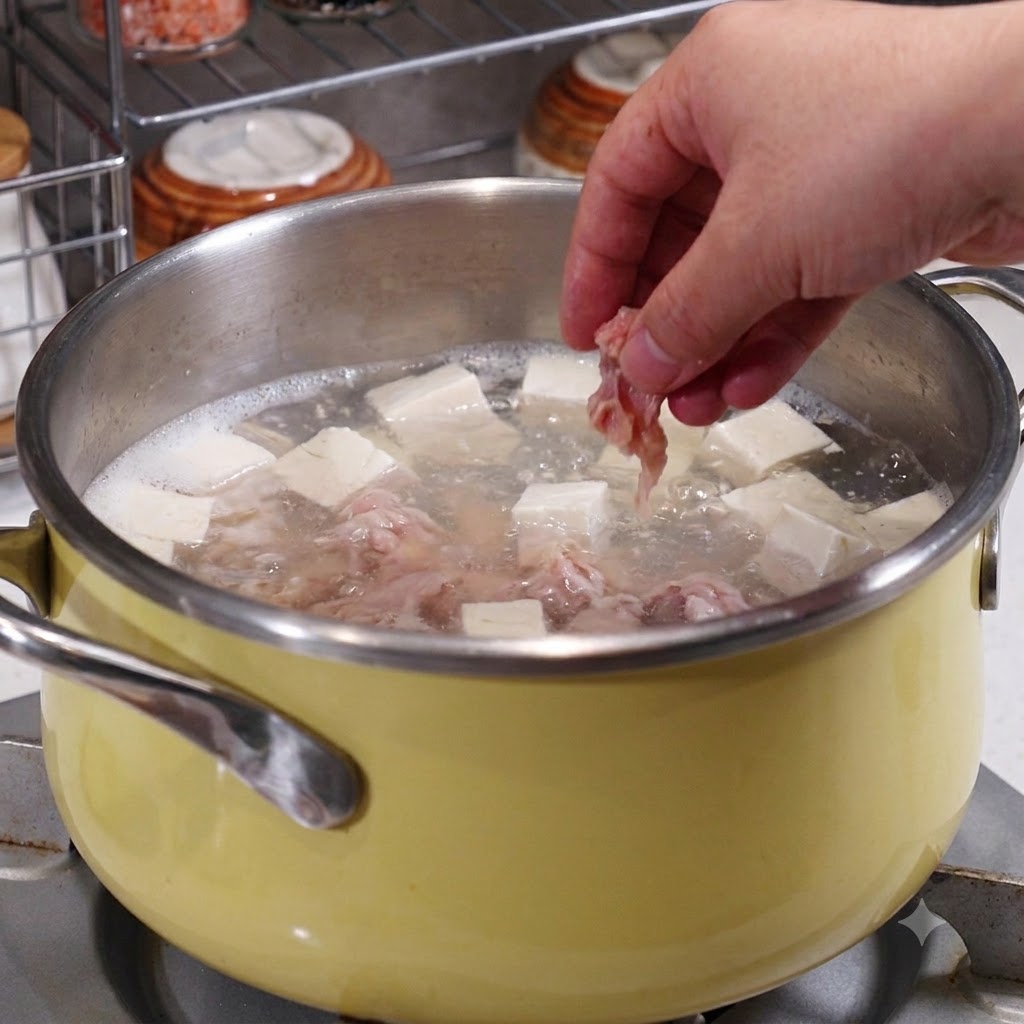 Una mano colocando rodajas de cerdo marinadas en una olla que contiene caldo hirviendo, tofu y mariscos.