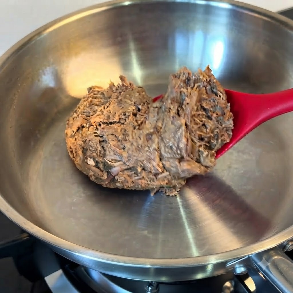 Un bloque de carne desmechada descongelada que se coloca en una sartén de acero inoxidable con una espátula roja.