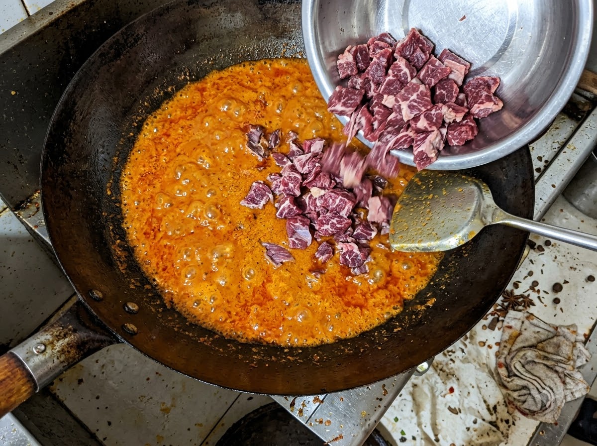 Trozos de falda de ternera siendo vertidos desde un bol metálico en una mezcla burbujeante de aceite de chile rojo dentro de un wok.
