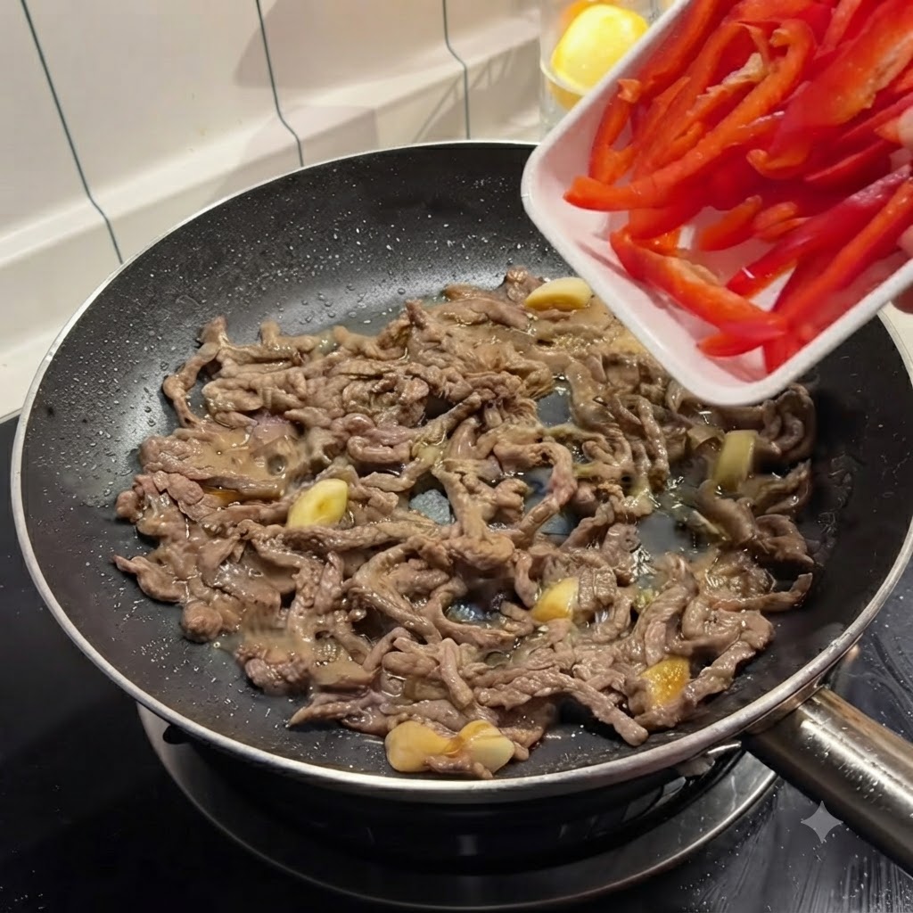 Tiras de pimiento rojo vertiéndose desde una bandeja blanca hacia una sartén con carne dorada.