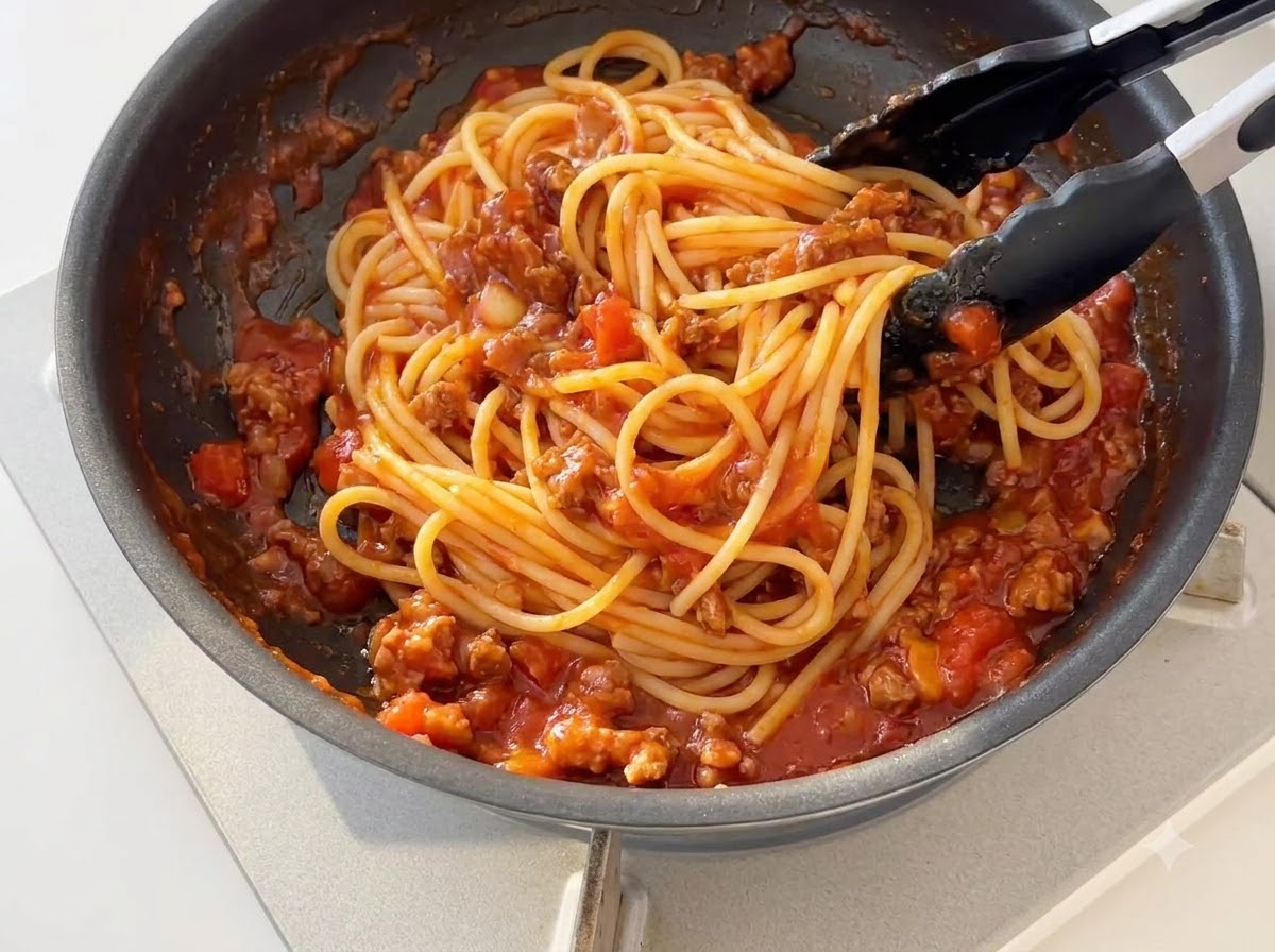 Fideos de espagueti cocidos siendo revueltos con salsa de tomate y carne espesa en una sartén usando pinzas de silicona negra.