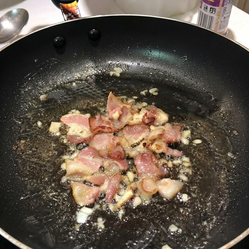 Pasta penne hervida mezclada con tocino y cebollas dentro de una sartén negra.