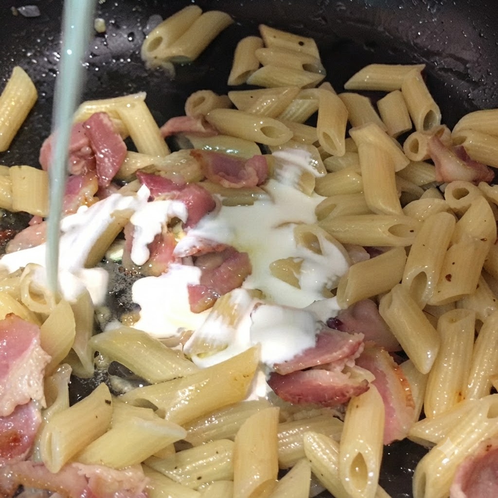 Leche y crema de leche siendo vertidas desde arriba en una sartén llena de pasta penne y trozos de tocino.