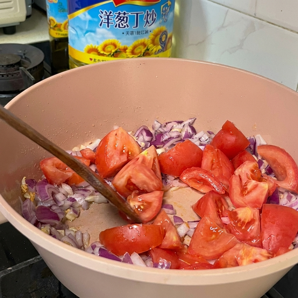 Trozos de tomates frescos mezclándose con cebollas moradas en cubitos y ajo en una sartén rosa usando una espátula de madera.