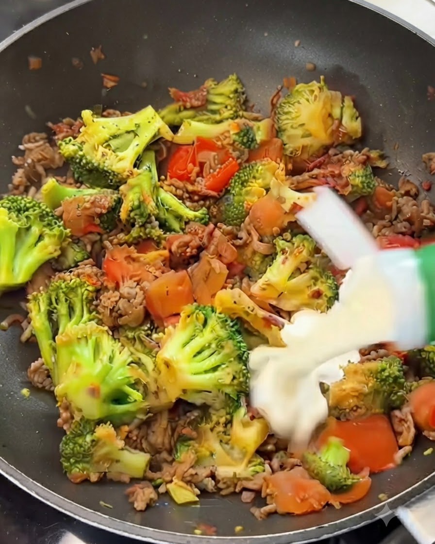 Crema de leche vertiéndose desde un envase sobre una mezcla de carne, brócoli y tomates en una sartén.