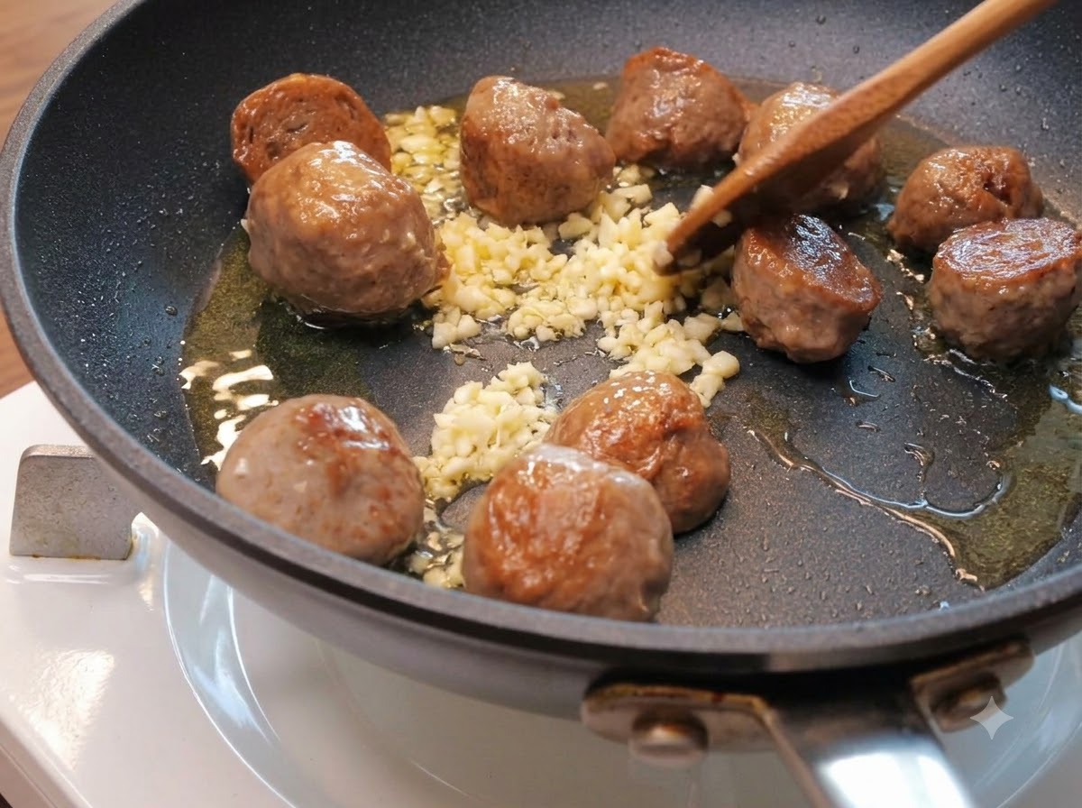 Ajo picado esparcido entre las mitades de albóndigas doradas en una sartén con aceite.