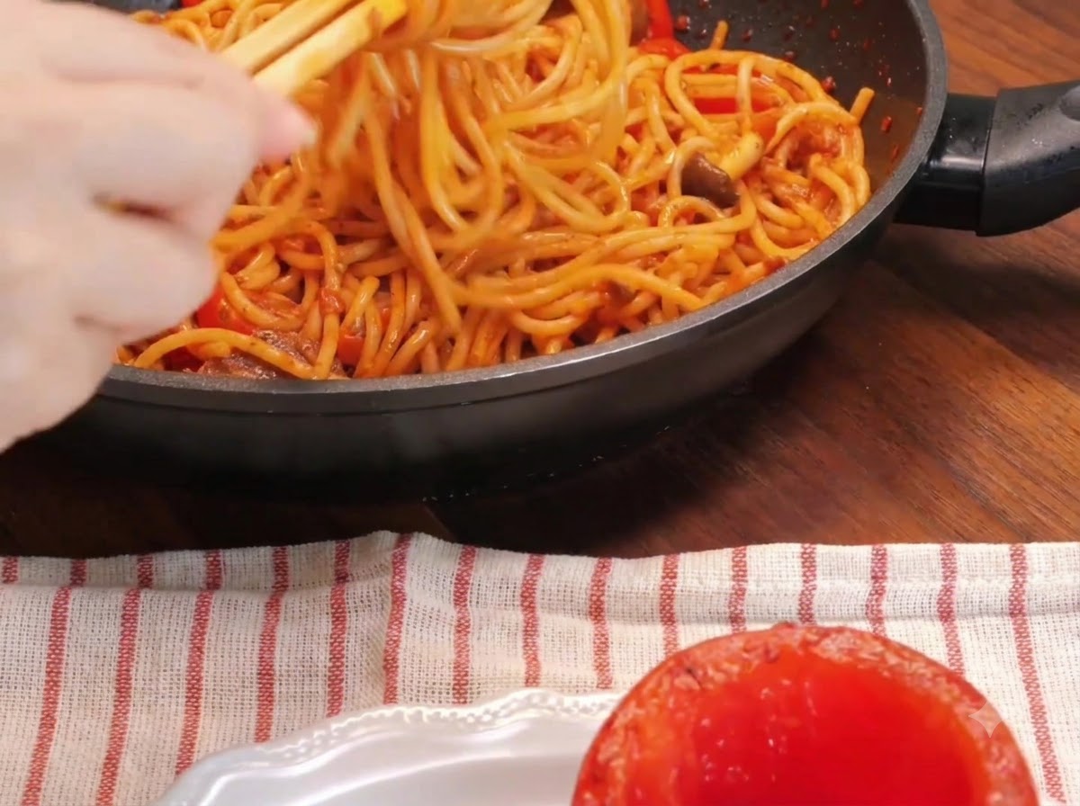 Un primer plano de una mano usando palillos para enrollar fideos de espagueti dentro de una sartén junto a un tomate asado ahuecado en un plato.