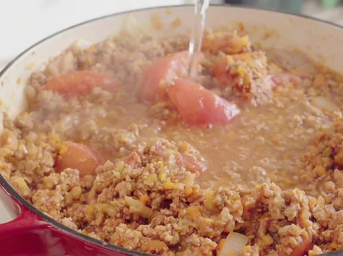 Agua vertida desde arriba en una olla roja grande que contiene la mezcla de carne, tomate y verduras a fuego lento.