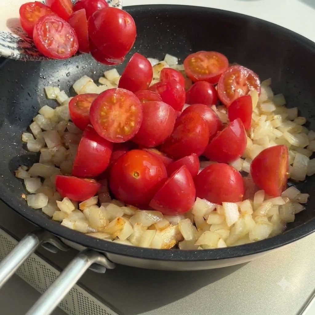 Mitades de tomates cherry agregadas a la sartén con cebolla y ajo, mezclándose juntos.