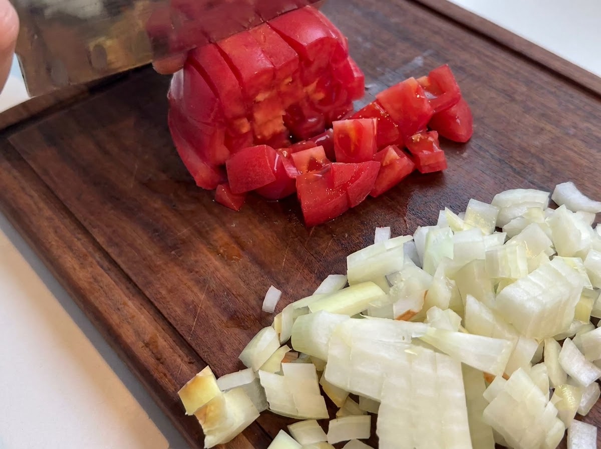 Tomate rojo fresco siendo cortado en cubitos con un cuchillo de cocina en una tabla de cortar de madera junto a cebollas picadas.
