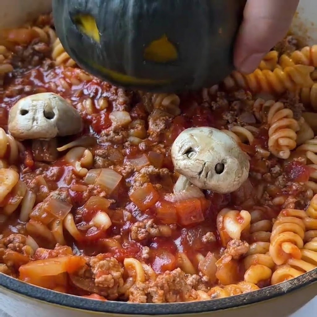 Una mini calabaza verde oscura tallada siendo bajada en el centro de una sartén llena de pasta de tomate cocida y salsa de carne picada.
