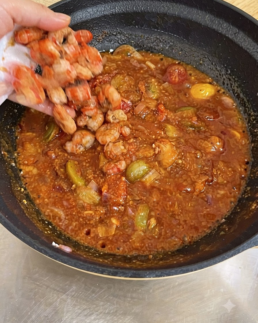 Una mano dejando caer camarones cocidos en una sartén llena de salsa de tomate roja burbujeante.