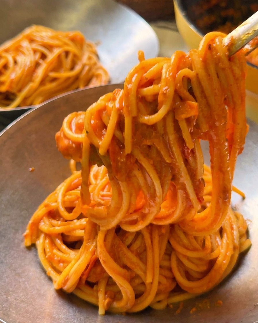 Pinzas levantando una porción de espagueti con tomate cremoso hacia un bol de servicio de metal pulido.