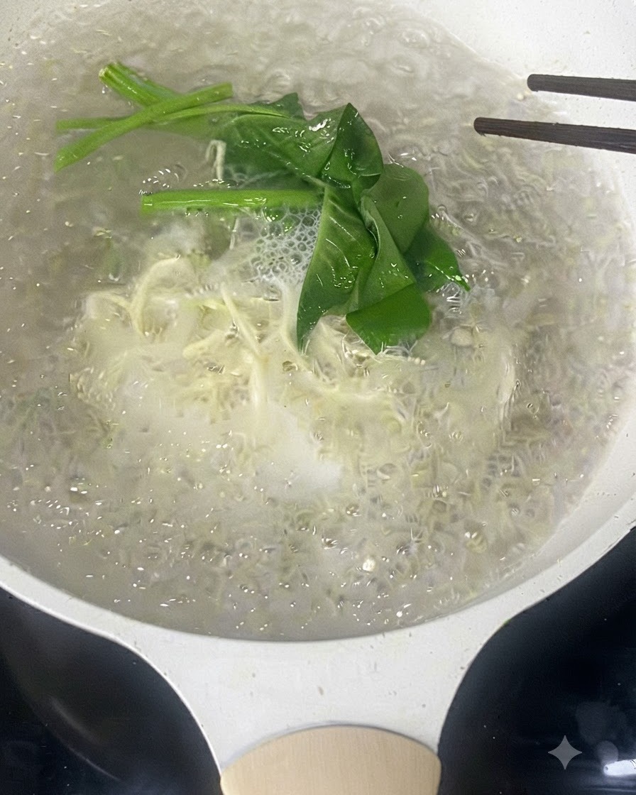Palillos removiendo fideos finos blancos y verduras de hoja verde hirviendo en agua.