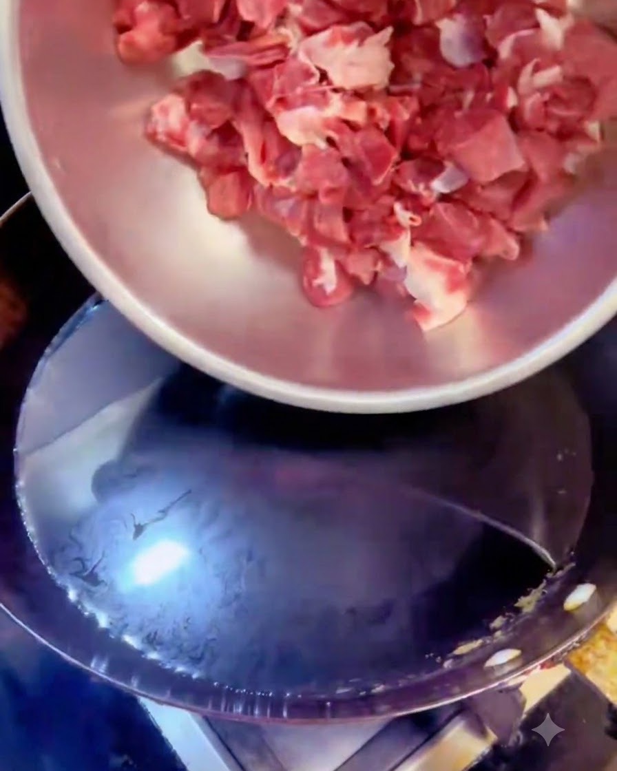 Cubos de carne siendo vertidos desde un bol de acero en una olla con agua hirviendo y aromáticos.