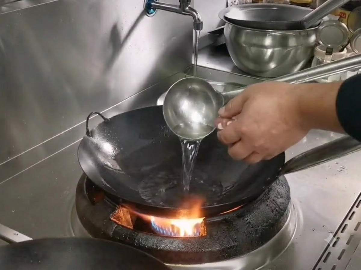 Un cucharón vertiendo aceite en un wok de hierro caliente sobre un quemador de gas mientras se prepara para cocinar cerdo desmenuzado.