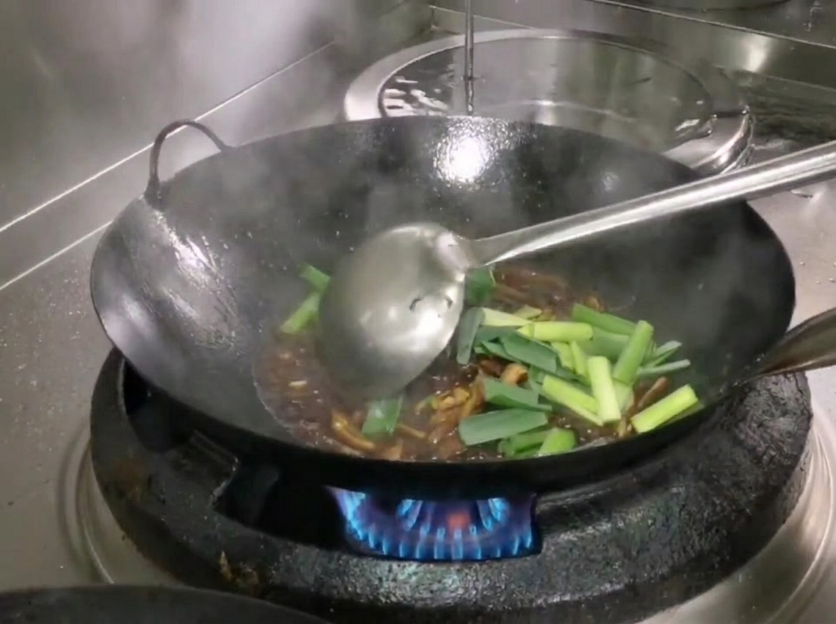 Secciones de brotes de ajo verde fresco añadidas al wok y mezcladas con los ingredientes usando un cucharón.