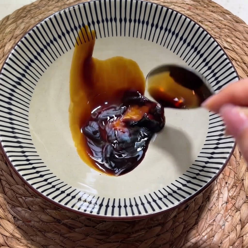 Una mano vertiendo salsa de soja oscura desde una cuchara en un cuenco de cerámica con motivos que contiene una cucharada de salsa de ostra.