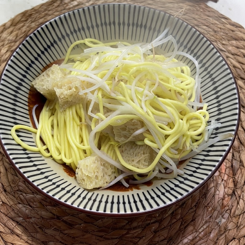 Fideos amarillos cocidos en un cuenco cubiertos con brotes de judía mungo pálidos y verduras encurtidas texturizadas.