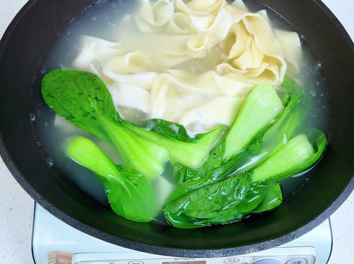 Palillos levantando los fideos anchos cocidos y el bok choy fuera del agua caliente.