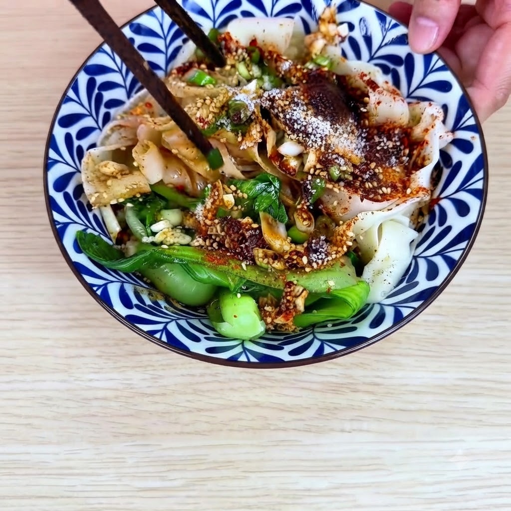 Palillos mezclando los fideos anchos de trigo, el bok choy y la salsa de aceite de chile juntos dentro del tazón de cerámica.