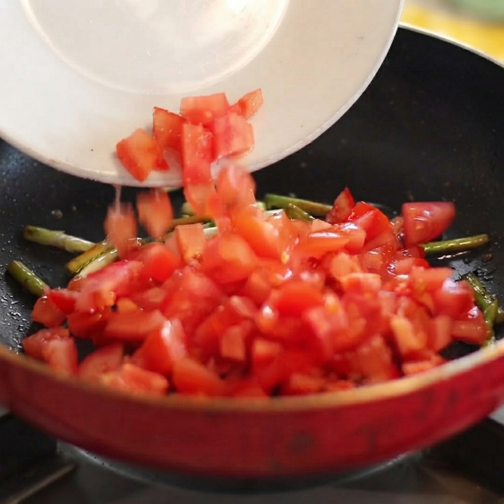 Tomates rojos en daditos deslizándose desde un plato blanco hacia una sartén caliente con cebolletas salteadas.