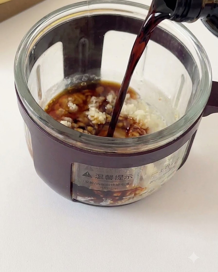 Salsa de soja oscura siendo vertida desde una botella en un recipiente de vidrio lleno de ajo picado y condimentos.