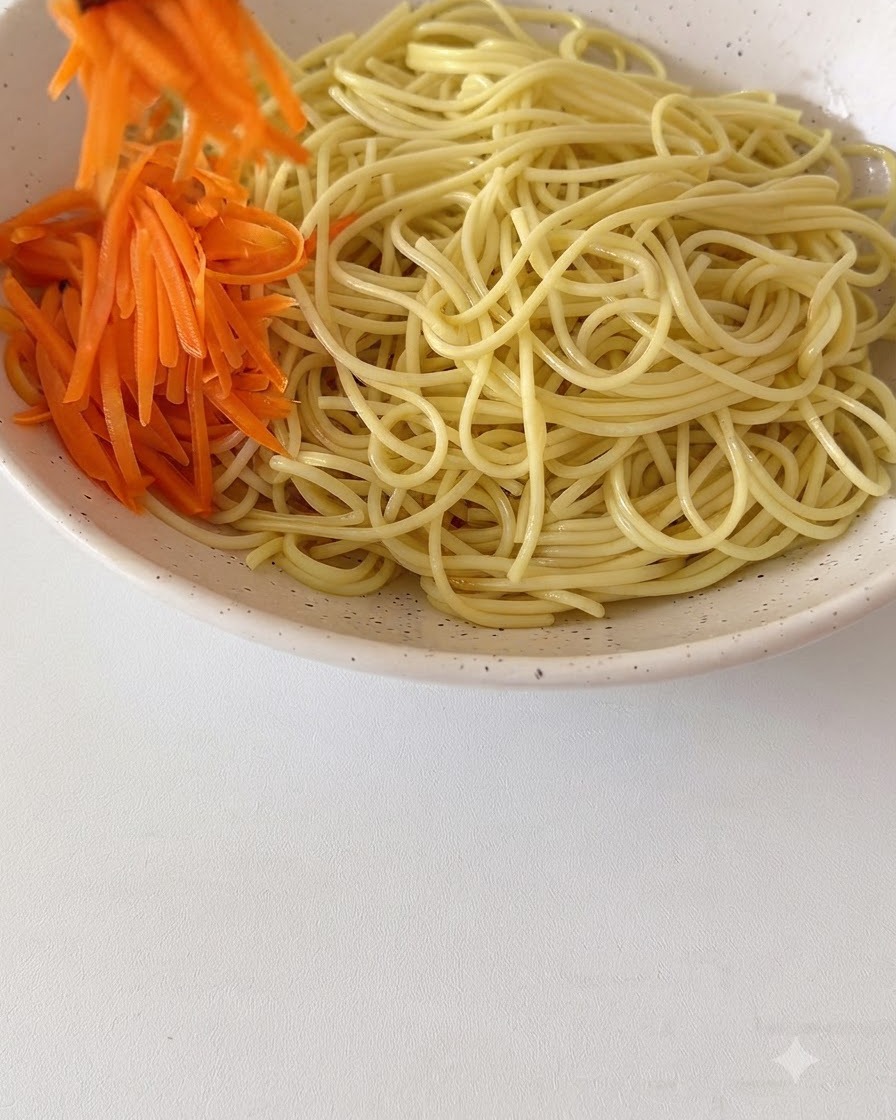 Zanahorias ralladas frescas y otros ingredientes siendo dispuestos encima de los fideos cocidos en un bol de cerámica grande.